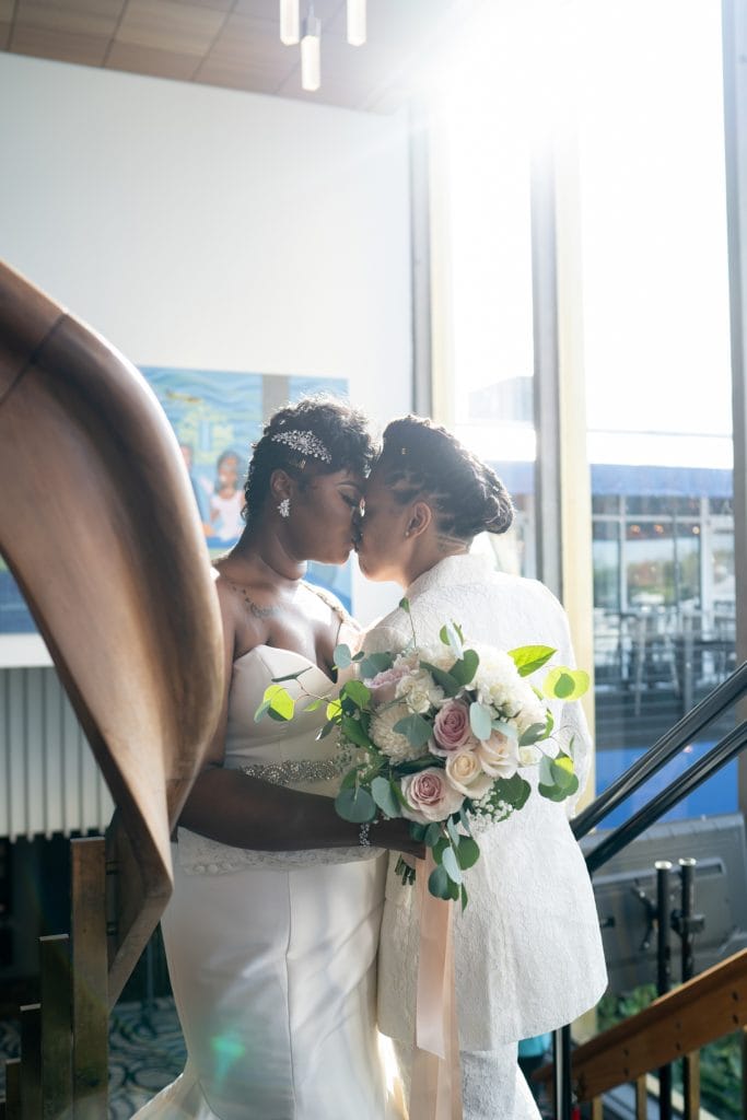 brides kissing on staircase with sunlight behind them.