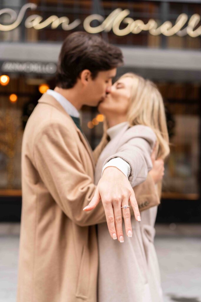 Newly engaged couple kissing while the woman holds her hand out to show off her ring.
